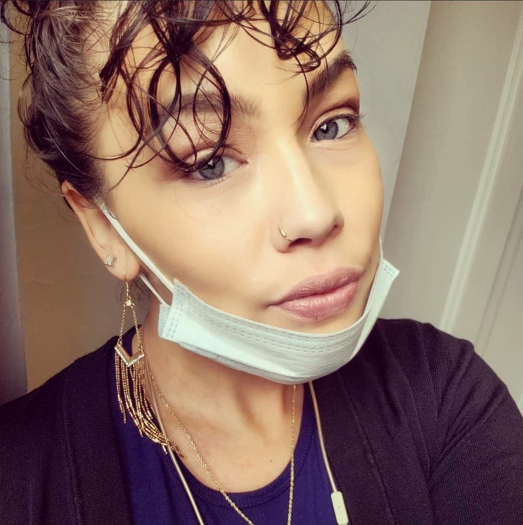 Woman with curly hair, face mask, and jewelry looking at the camera.