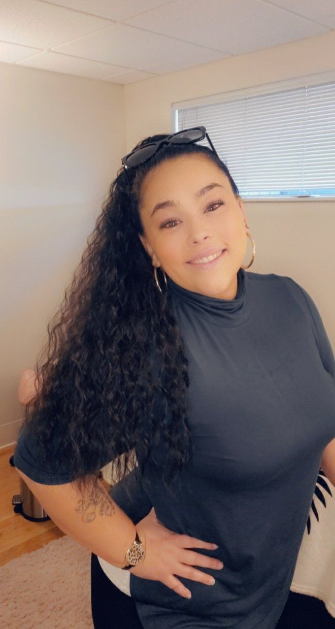Woman with dark curly hair, smiling, hand on hip, wearing a black top and gold hoop earrings.