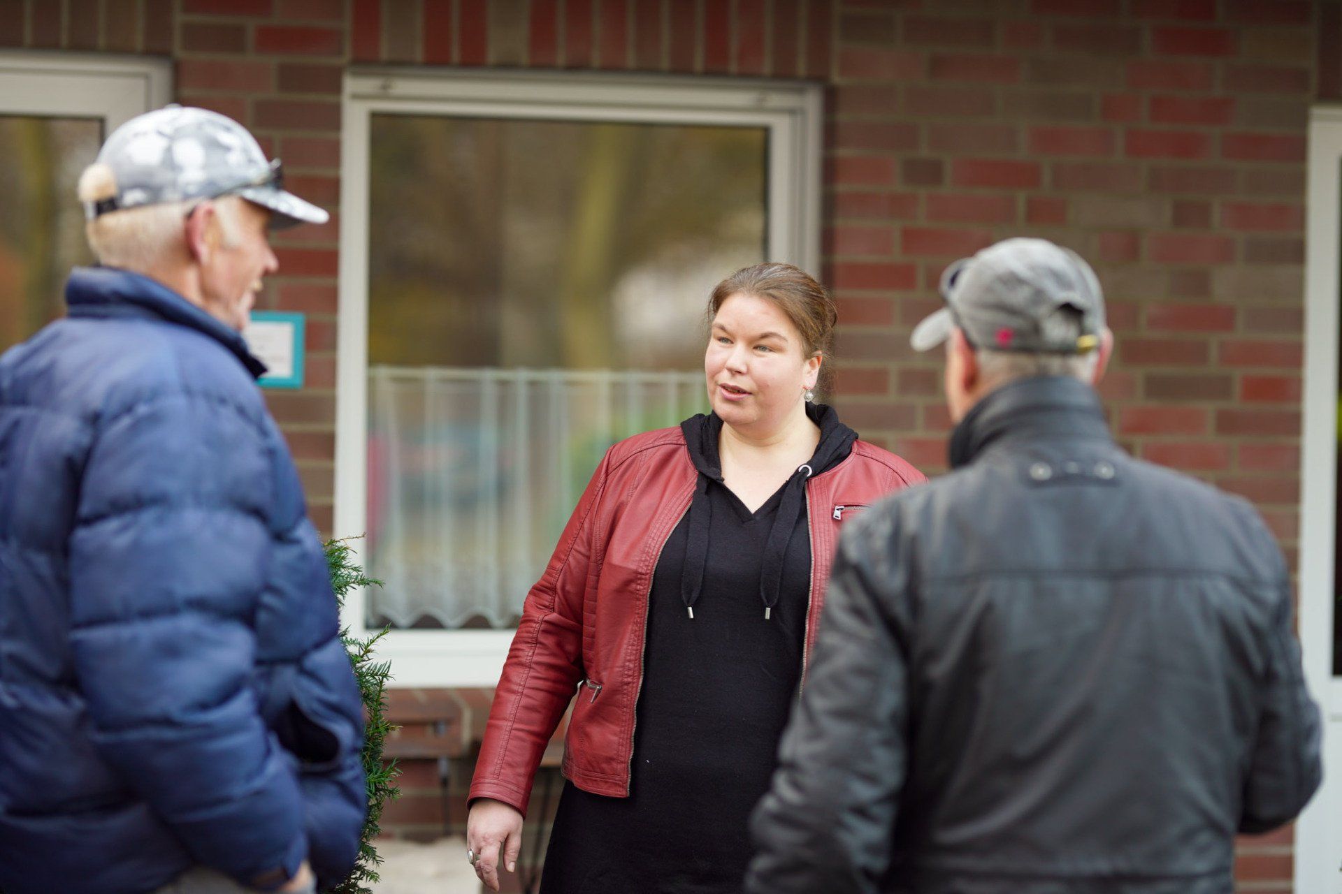 Eine Frau in einer roten Jacke spricht mit zwei Männern vor einem Gebäude.
