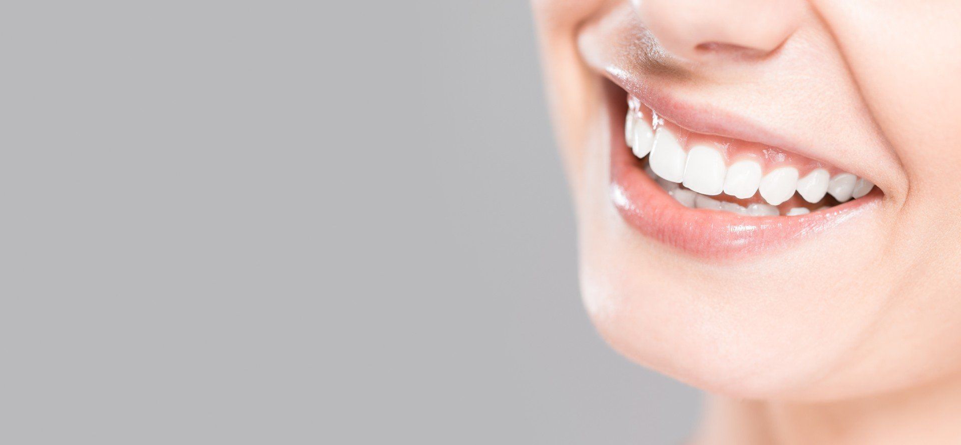 Close-up of a person's smiling mouth, showing bright white teeth, pink lips, and a light gray background.