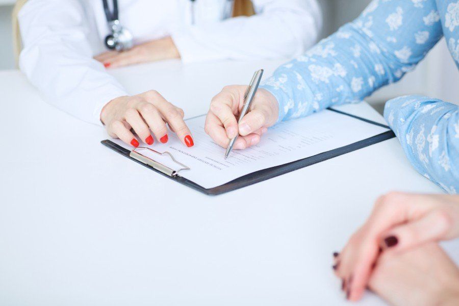 Doctor writing on a clipboard, pointing to a form while consulting with a patient at a desk.