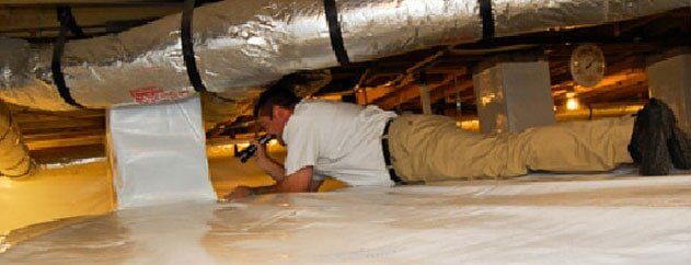 Person crawling in a low crawlspace beneath silver ductwork, examining insulation.