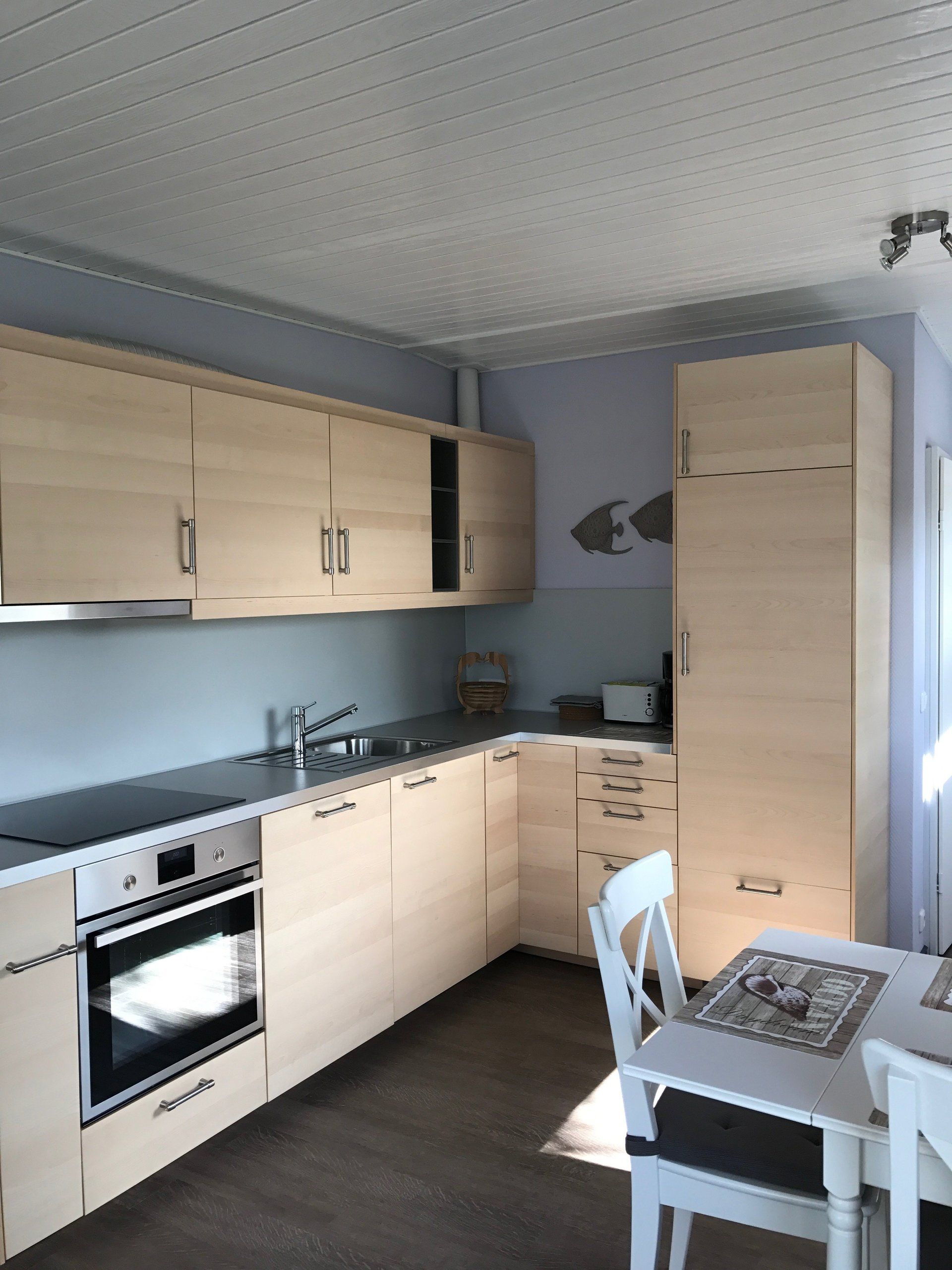 Eine Küche aus hellem Holz mit Geräten, Schränken und einem Tisch. Hellblaue Wände, weiße Decke.