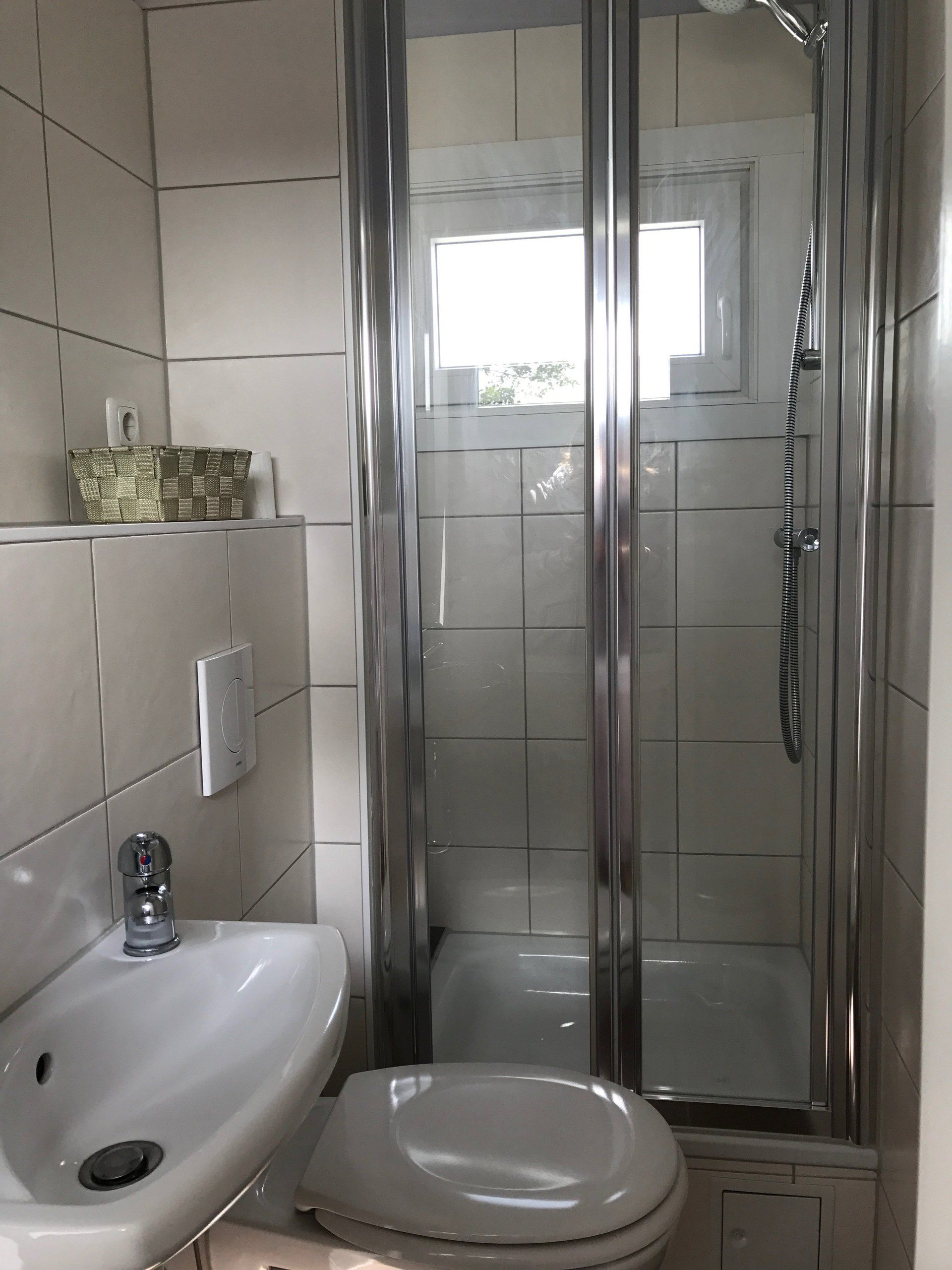 Kleines Badezimmer mit Dusche, Toilette und Waschbecken. Weiße Fliesen, silberner Duschtürrahmen, kleines Fenster.