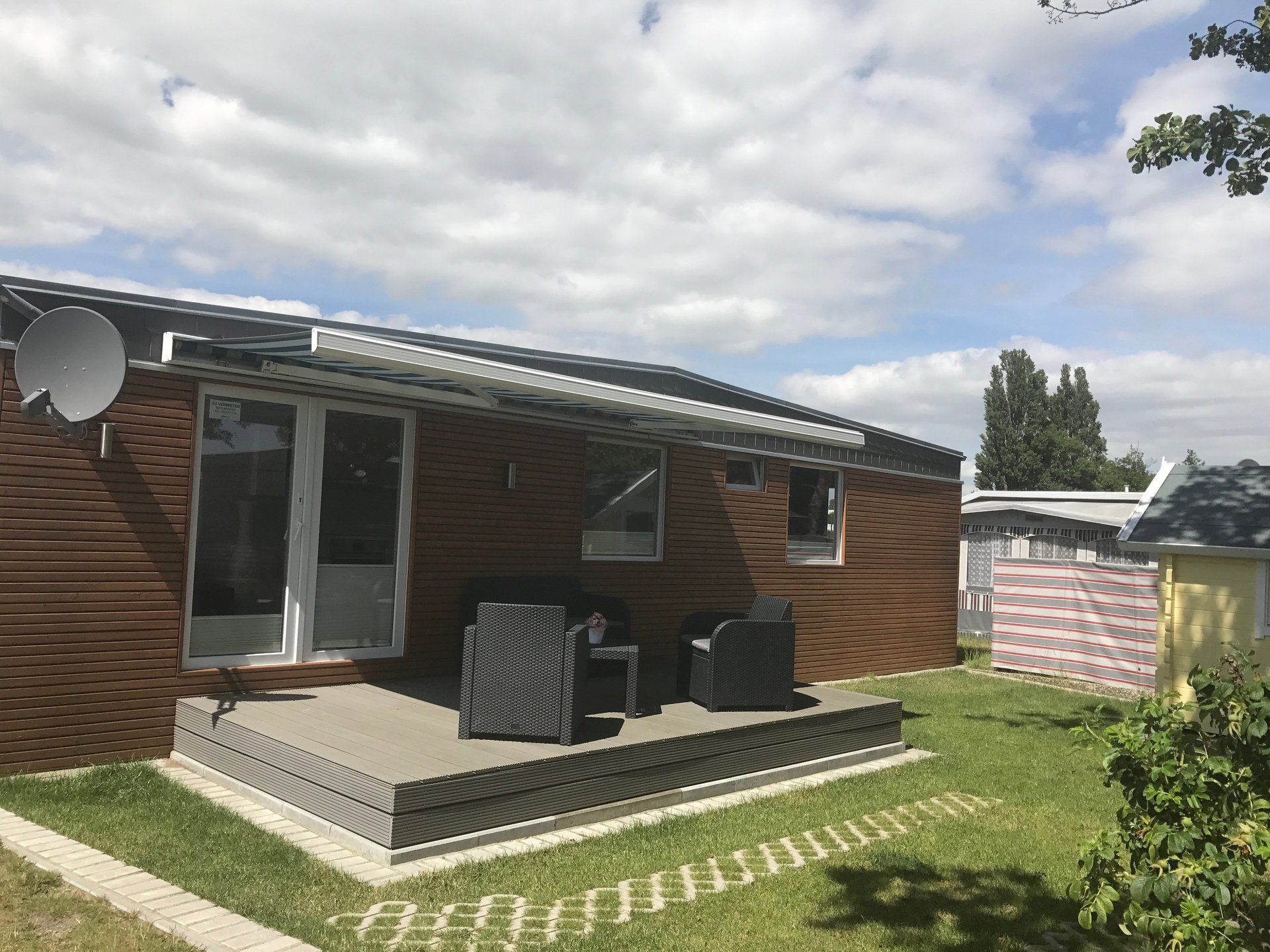 Braune Hütte mit Terrasse, Gartenmöbeln und Satellitenschüssel unter einem bewölkten Himmel.
