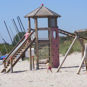 Kinder spielen auf einem Holzspielplatz mit Rutsche und Klettergerüst an einem Sandstrand.