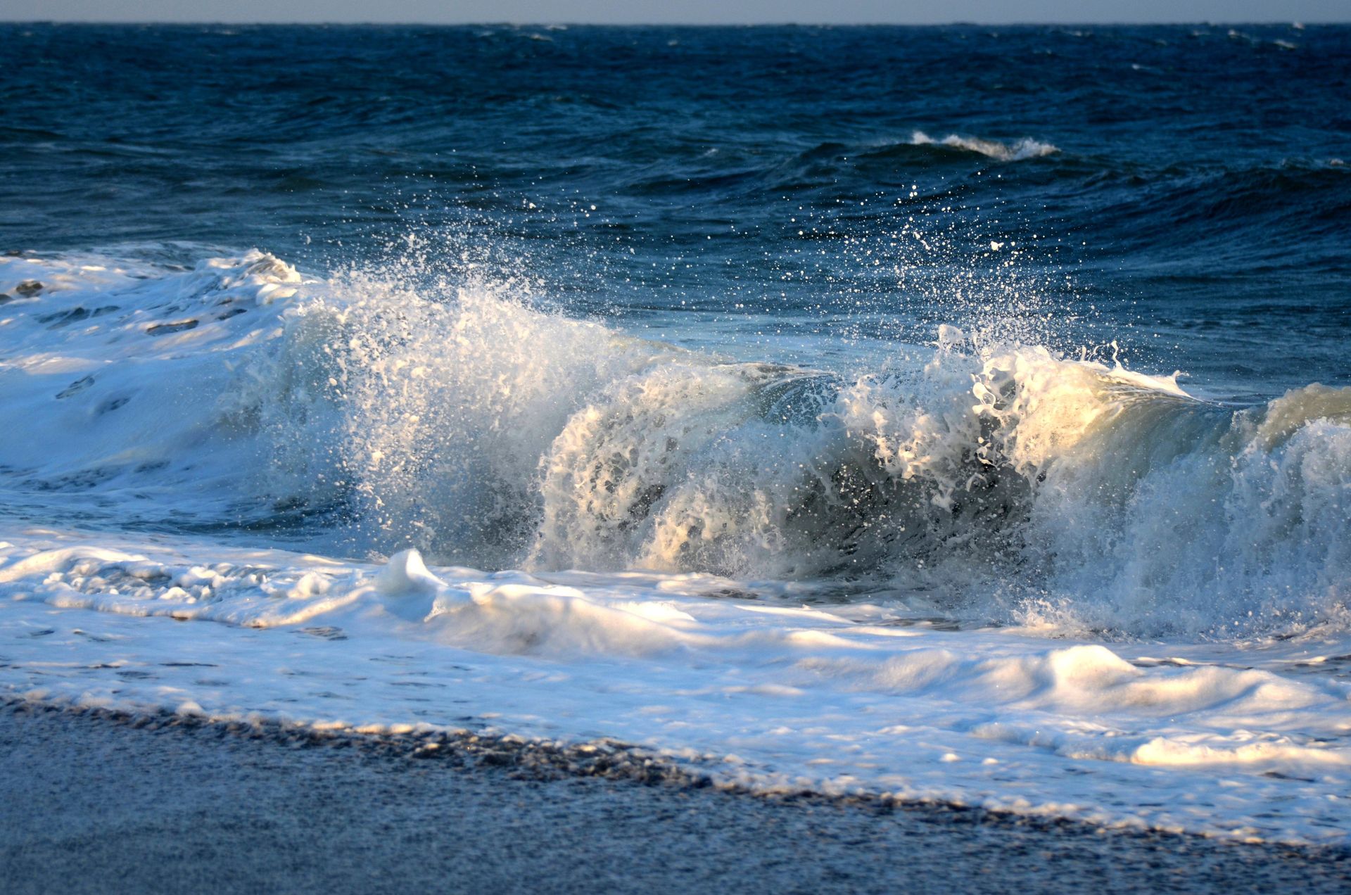 Brechende Meereswelle an einem Strand mit weißem Schaum und plätscherndem Wasser unter blauem Himmel.