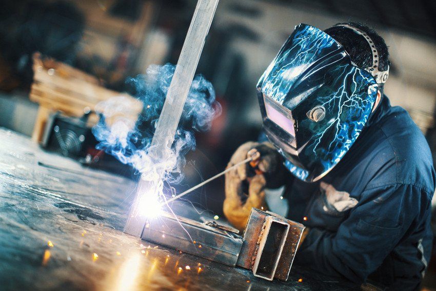 Closeup side view of unrecognizable worker welding two metal plates with gas welding machine.He's wearing welding mask and protective workwear and gloves.