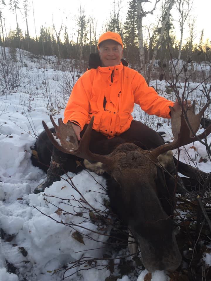 A man in an orange jacket is sitting next to a moose in the snow.