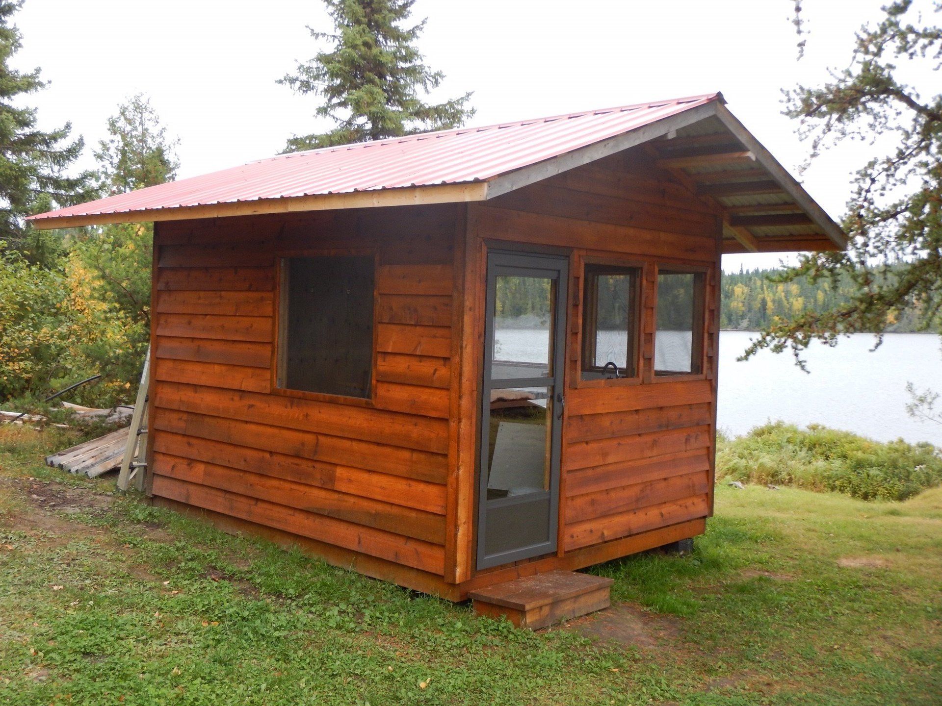 A small wooden cabin with a red roof sits in the grass near a lake