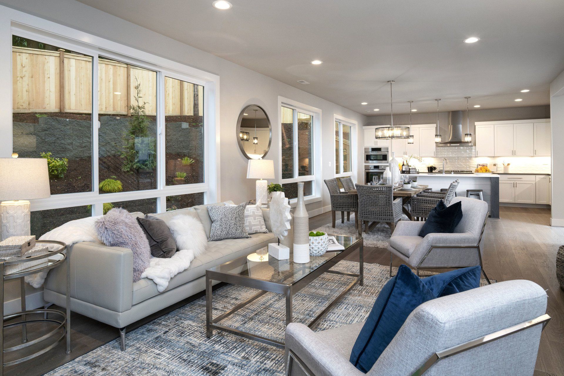 Bright modern living room with gray sofas, blue chair, glass coffee table, and open kitchen beyond.