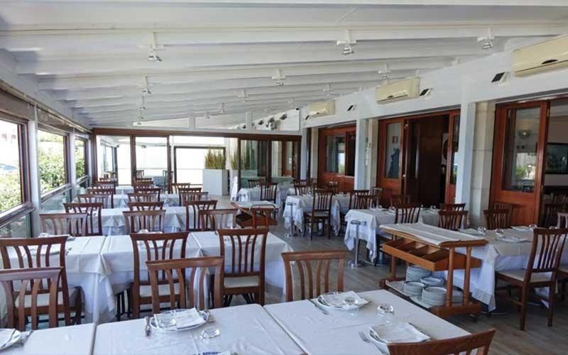 Sala da pranzo vuota di un ristorante con tovaglie bianche, sedie di legno e grandi finestre lungo la parete sinistra.