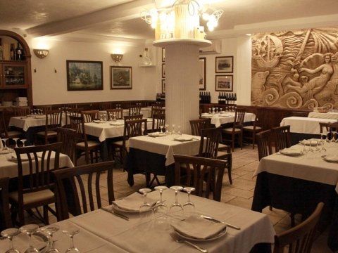 Sala da pranzo vuota di un ristorante con tavoli apparecchiati, sedie in legno scuro e luci calde alle pareti.