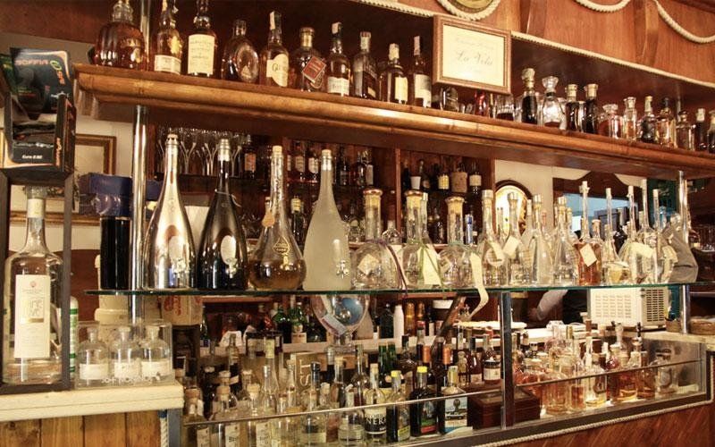 Dietro un bancone di legno si trovano scaffali pieni di bottiglie di liquore e bicchieri di vario tipo.