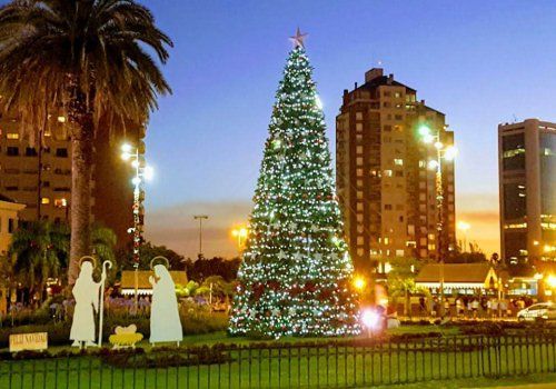 Árbol de Navidad iluminado en una plaza de la ciudad al anochecer, con figuras del nacimiento y edificios al fondo.
