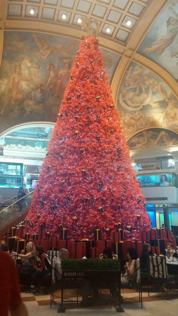Un alto árbol de Navidad rojo en un gran atrio interior con techo ornamentado y gente debajo.