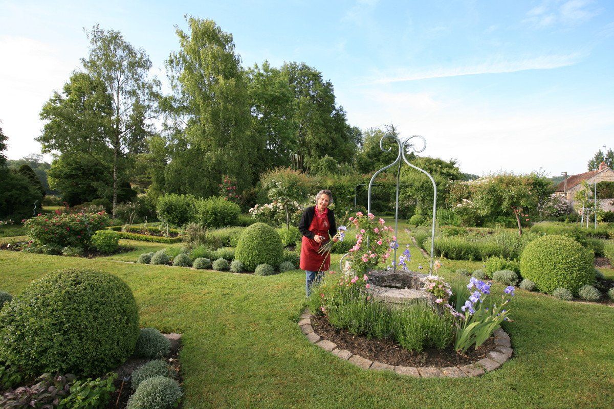 Bertrande Deladoucette se tient dans un jardin luxuriant, près d'un parterre de fleurs et d'une treille blanche.