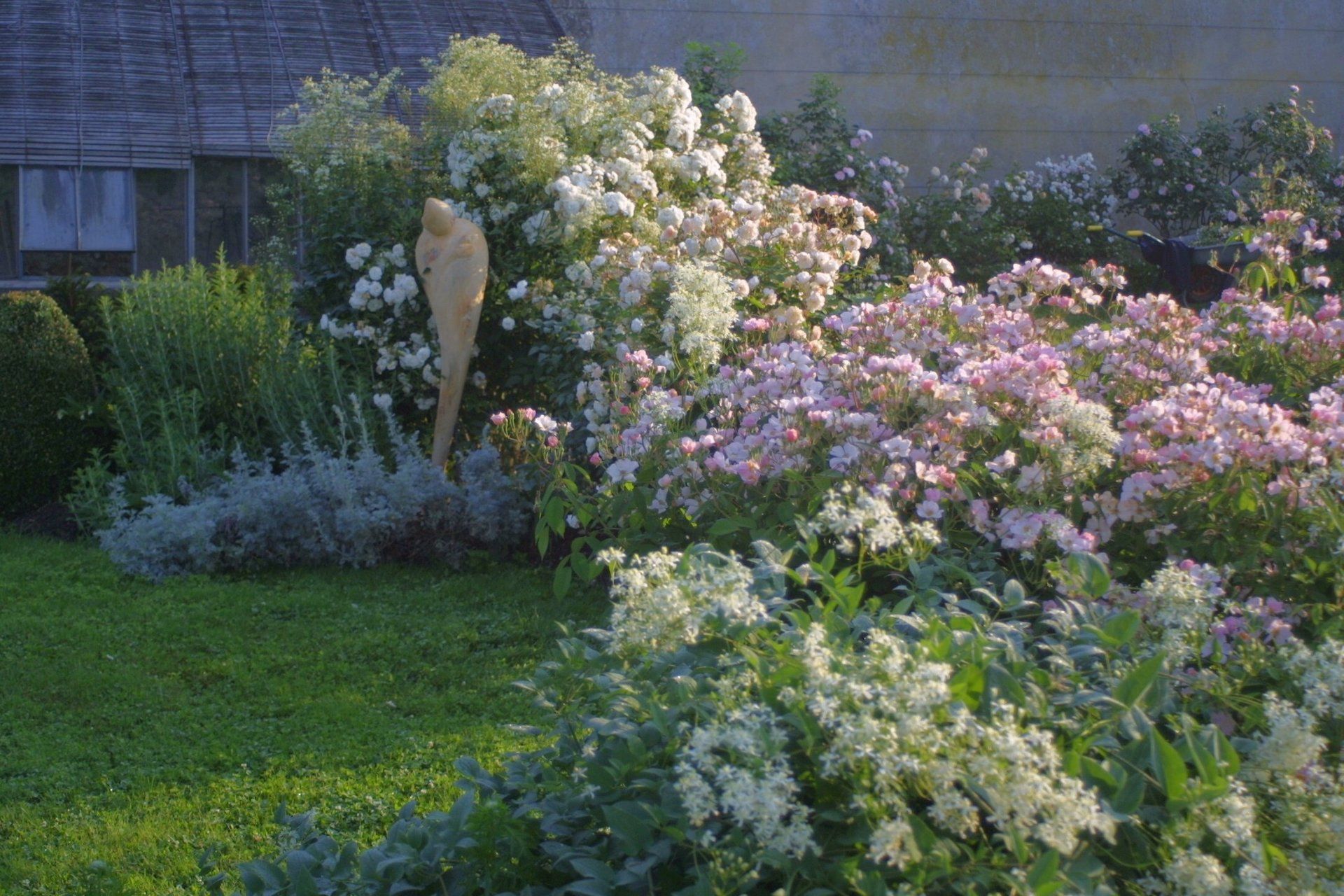 Jardin luxuriant avec des fleurs roses, blanches et jaunes épanouies, bordant une pelouse verte et une maison.