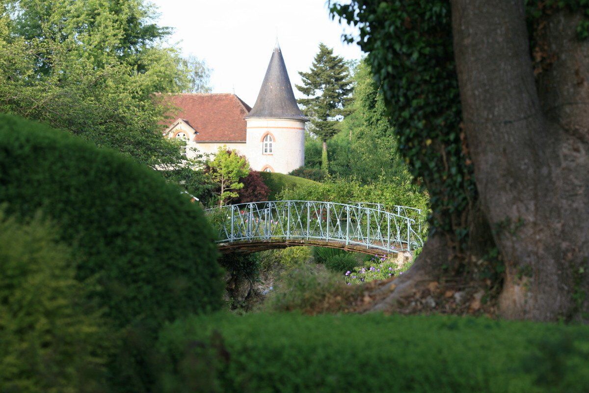 Passerelle dans un jardin luxuriant menant à une petite tour de château au toit rouge et à la tourelle conique.