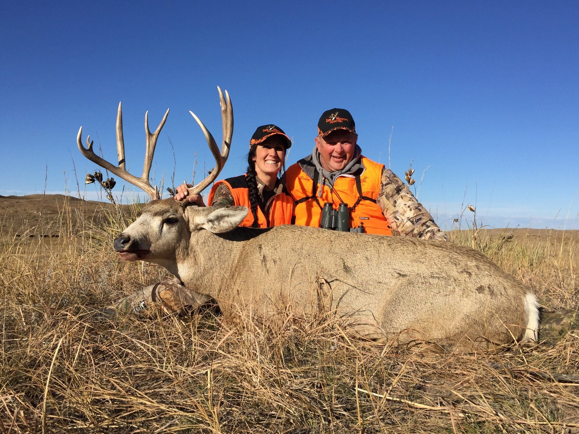 Two hunters pose with a large buck in tall grass. The sky is blue and they wear orange vests and hats.