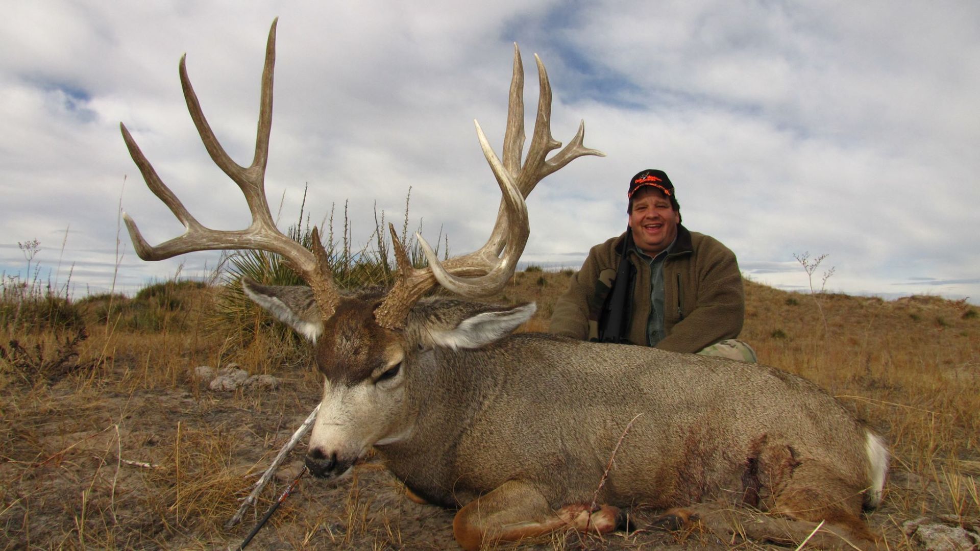 Hunter kneels next to a large mule deer buck with massive antlers in a field under a cloudy sky.