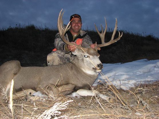 Hunter kneeling, holding large buck with impressive antlers, outdoors. Dusk setting, hunter smiling.