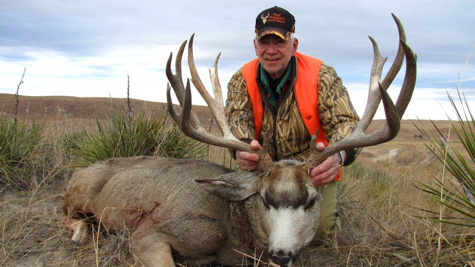 Man kneels beside a large deer, holding its massive antlers. He wears camo and orange vest; outdoor setting.