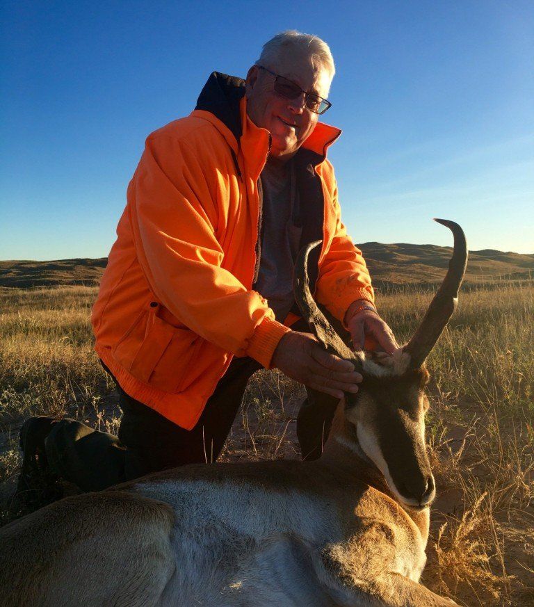 Man in orange jacket kneels beside an antelope in a grassy field; the man smiles and touches the animal.