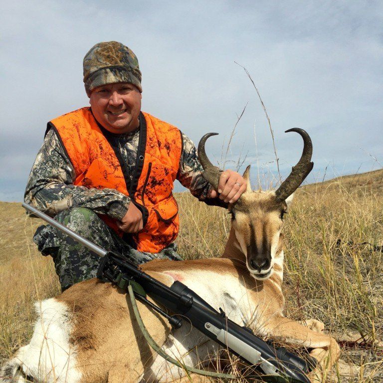 Man in orange vest kneels next to a harvested pronghorn antelope on a grassy hillside.