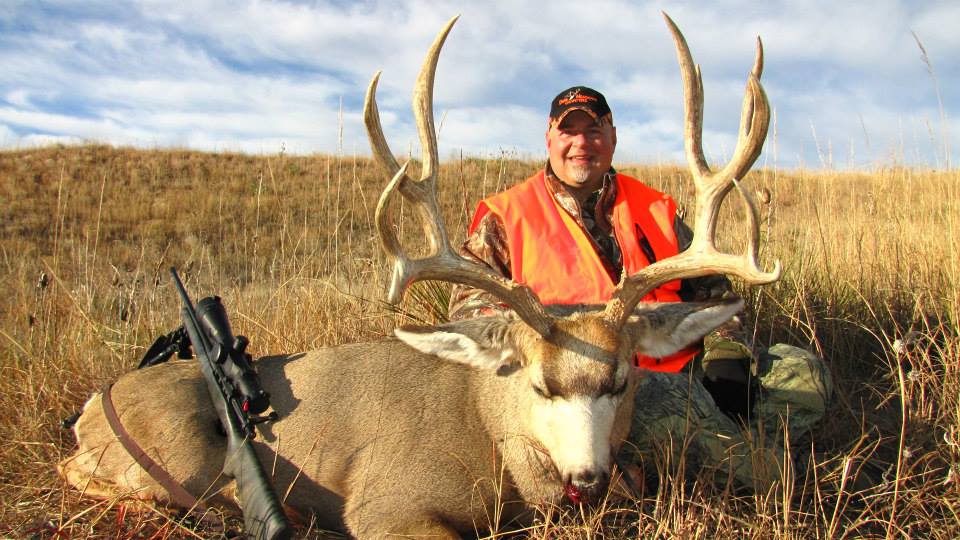 Man in orange vest kneels beside a large deer with large antlers in a grassy field.