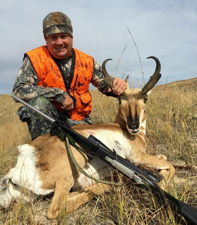 Hunter kneels beside a pronghorn antelope on a grassy hill. He wears orange vest and cap, smiles, and holds the antelope's horns.
