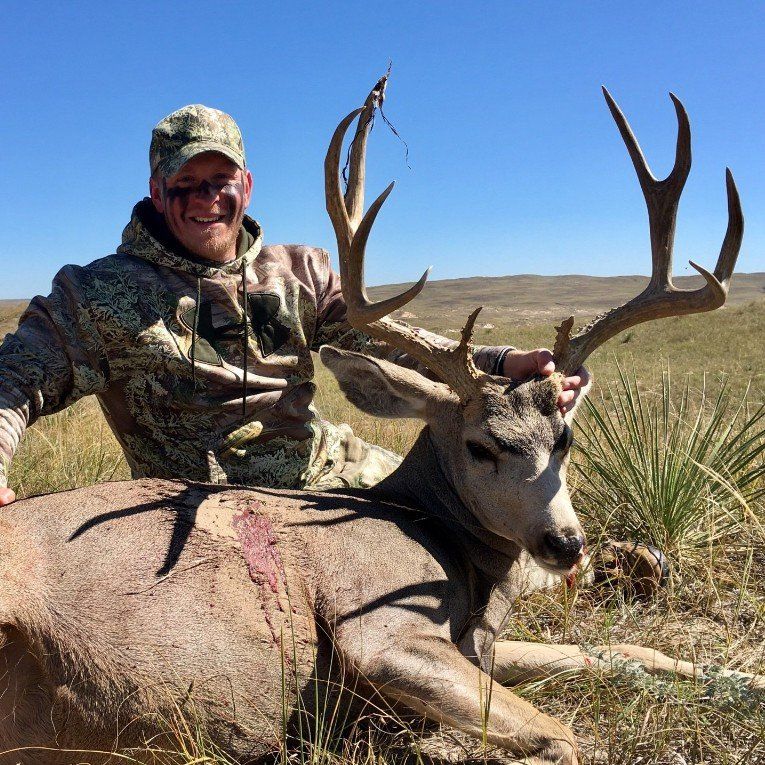 Man in camouflage smiles, holding a large buck with impressive antlers in a grassy field.