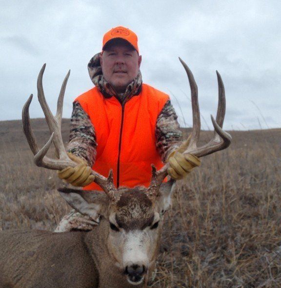 Man in orange vest and hat holds a large buck with impressive antlers, posing outdoors.