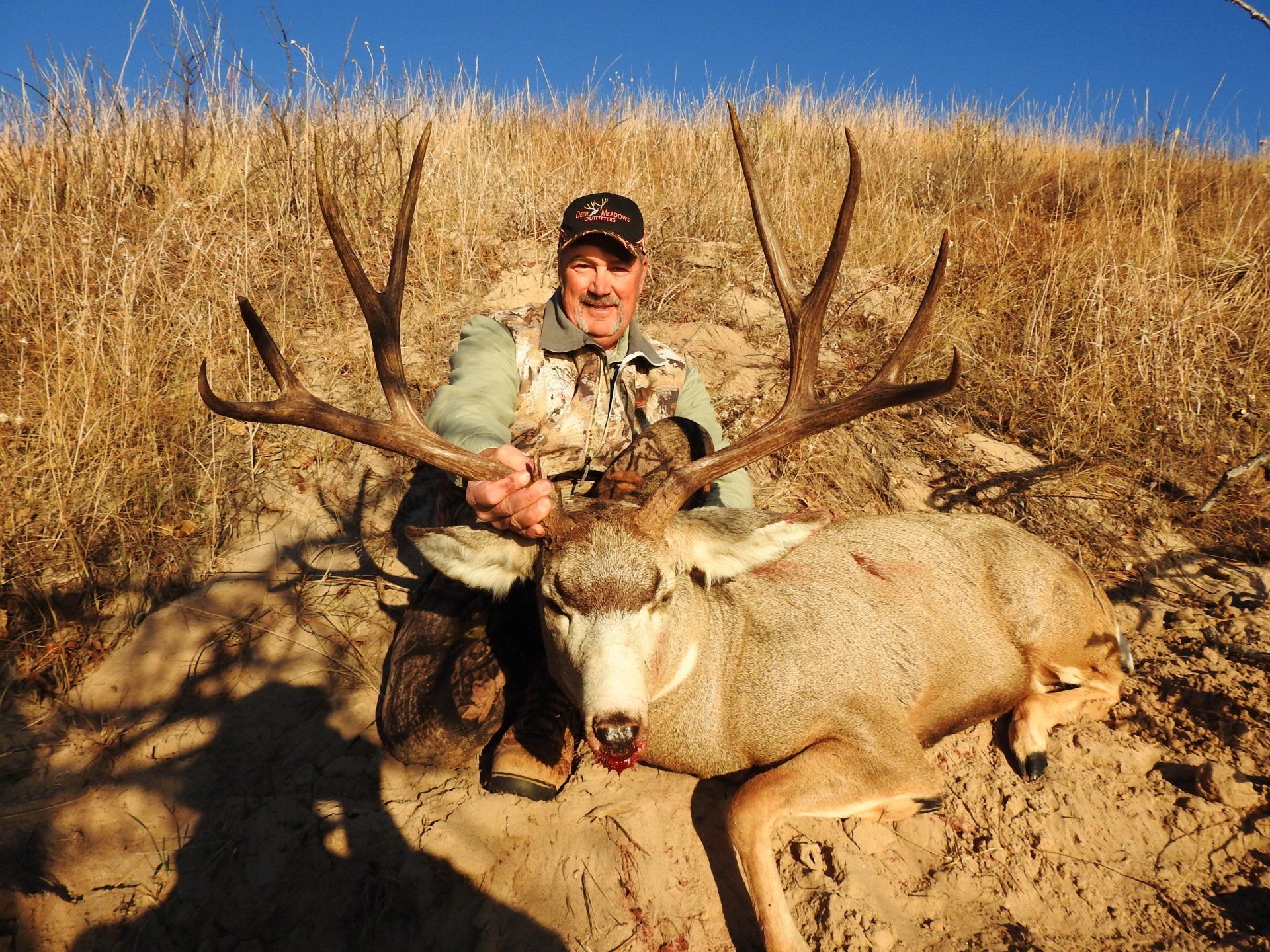Man kneeling with large, dead deer with impressive antlers in a dry, grassy field under a blue sky.