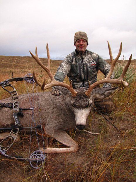 Man in camo with a large buck he harvested, bow beside it. Setting is a grassy field.