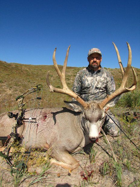 Man in camouflage kneels next to a large deer with impressive antlers on a grassy hillside.