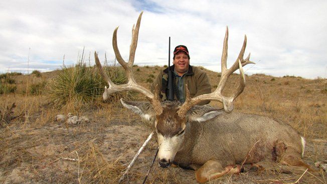 Hunter with a large deer, holding rifle in a grassy setting. Deer has large antlers, hunter is smiling.