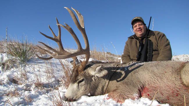 Man smiles next to a large deer lying in the snow, rifle in hand. Blue sky, brown coat.