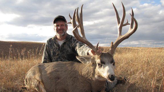 Man in camouflage smiles, poses with large buck in field.