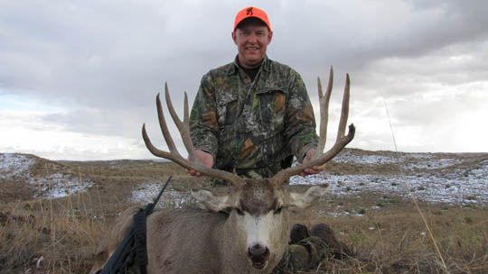 Hunter in orange hat, smiling, holds large mule deer buck with impressive antlers in a field.