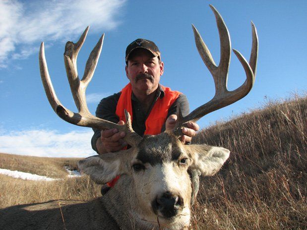 Man holding a large deer with antlers in a field; he wears an orange vest and a hat.