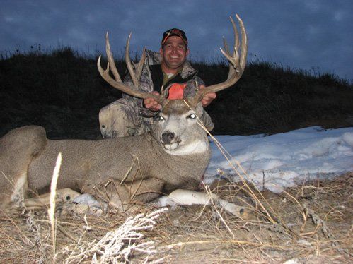 Man in camo holds up large buck antlers, smiles. The buck lies on snow and grass outdoors at dusk.