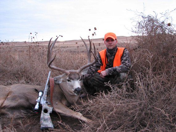 Hunter kneels beside a large buck he harvested. Field setting, cloudy sky. Hunter wears orange vest and hat.