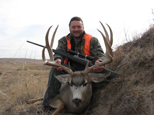 Man in orange vest, smiling, kneels beside a harvested buck with large antlers, holding rifle. Outdoors on a grassy hill.