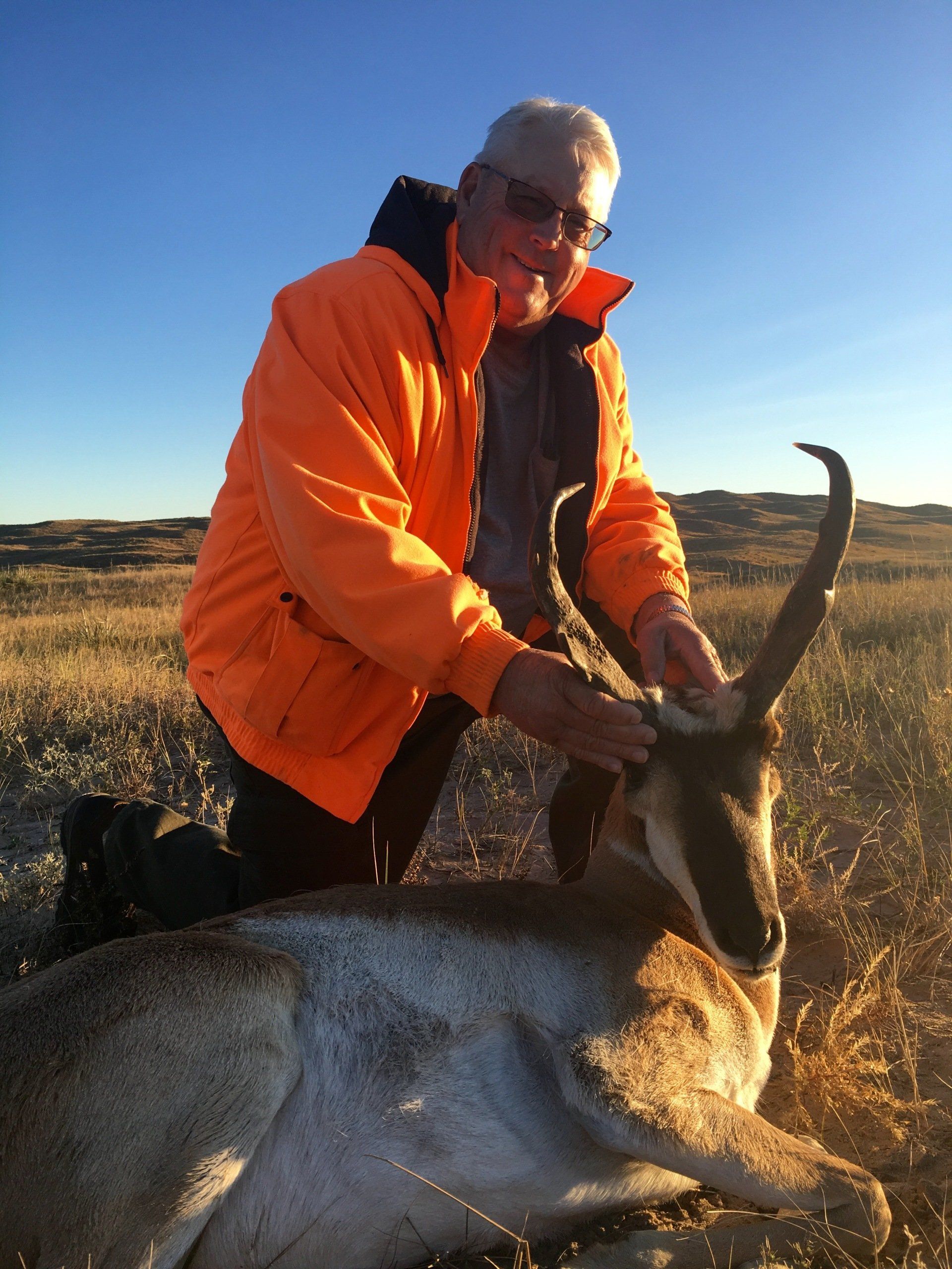 Hunter kneels next to a pronghorn antelope. He's wearing orange, outdoors on a sunny day.