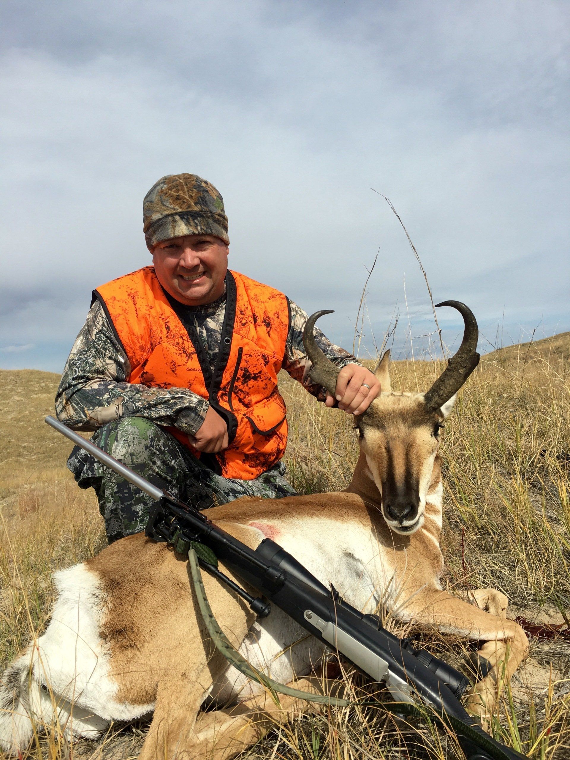 Man in orange vest kneels beside a dead pronghorn on a grassy hillside, rifle present.