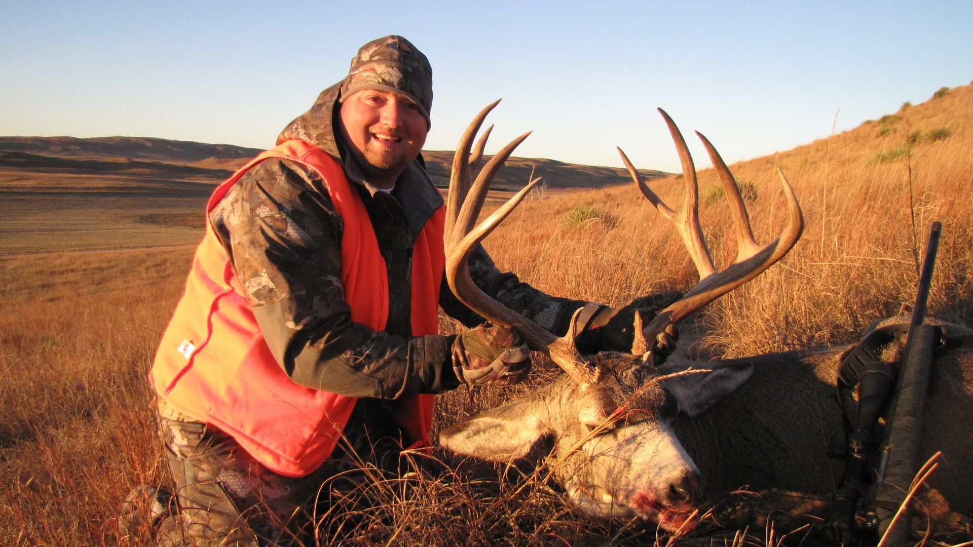 Hunter in orange vest smiles next to a harvested buck with large antlers in a grassy field.
