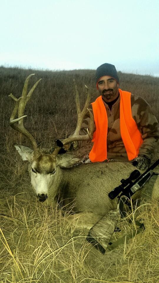 Hunter in orange vest smiles next to a large buck with impressive antlers in a grassy field.