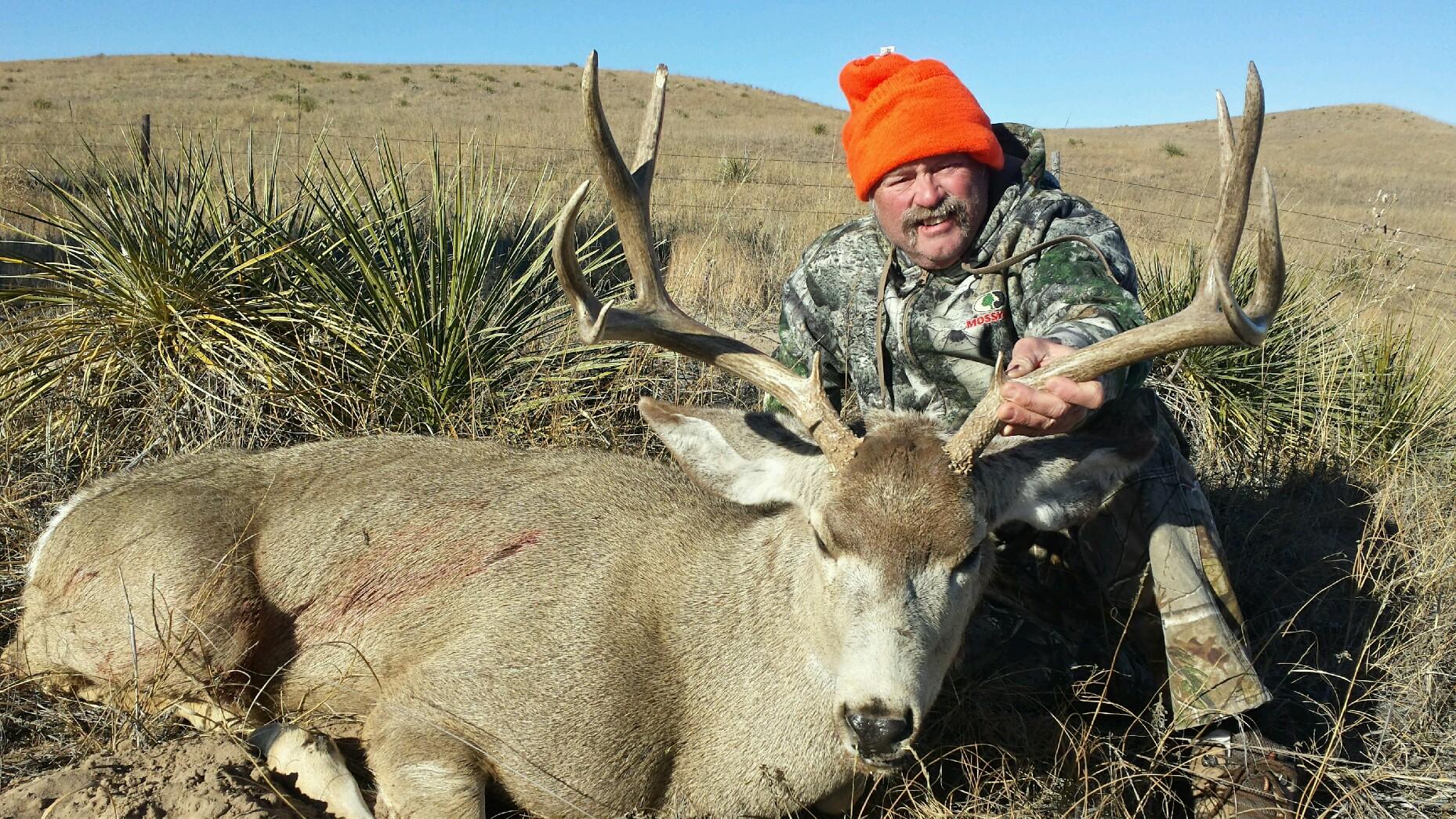 Hunter in orange hat poses with a large dead deer in a desert landscape.
