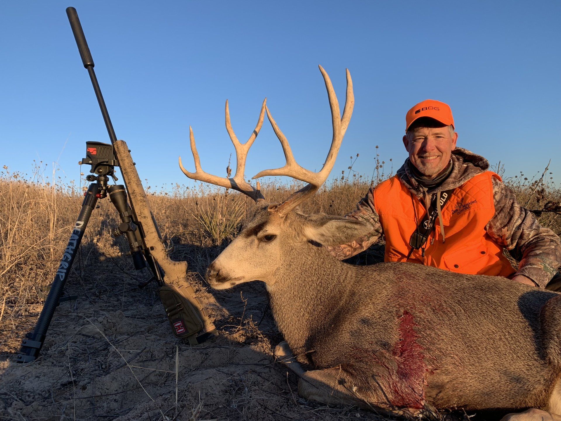 Hunter kneels beside a large buck, rifle nearby. Sunny day, orange vest, field.